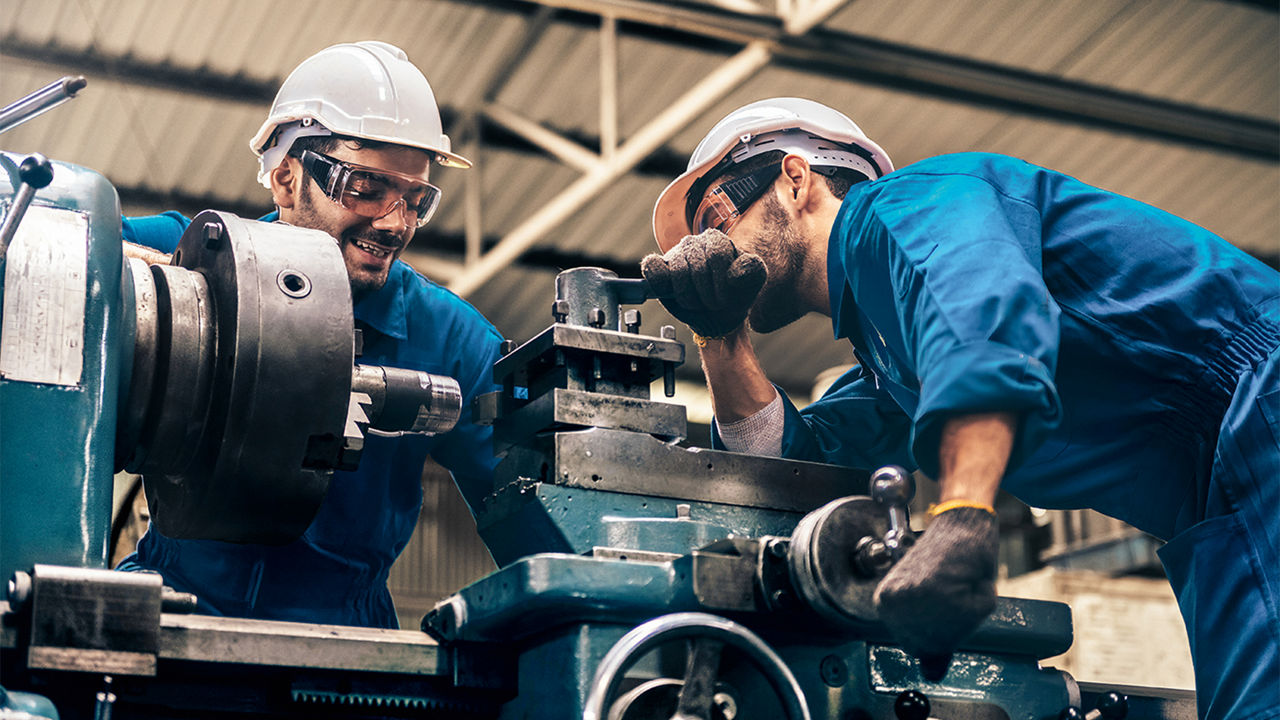 Zwei Männer mit Blaumann, Helm und Schutzbrille arbeiten in einer Werkstatt an einer Drehbank