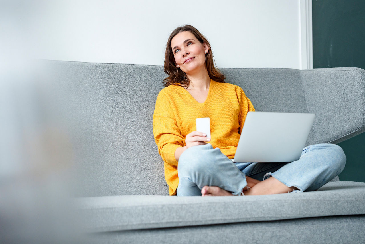 Eine Frau sitzt im Schneidersitz auf einer grauen Couch mit einem aufgeklappten Laptop auf ihrem Schoß