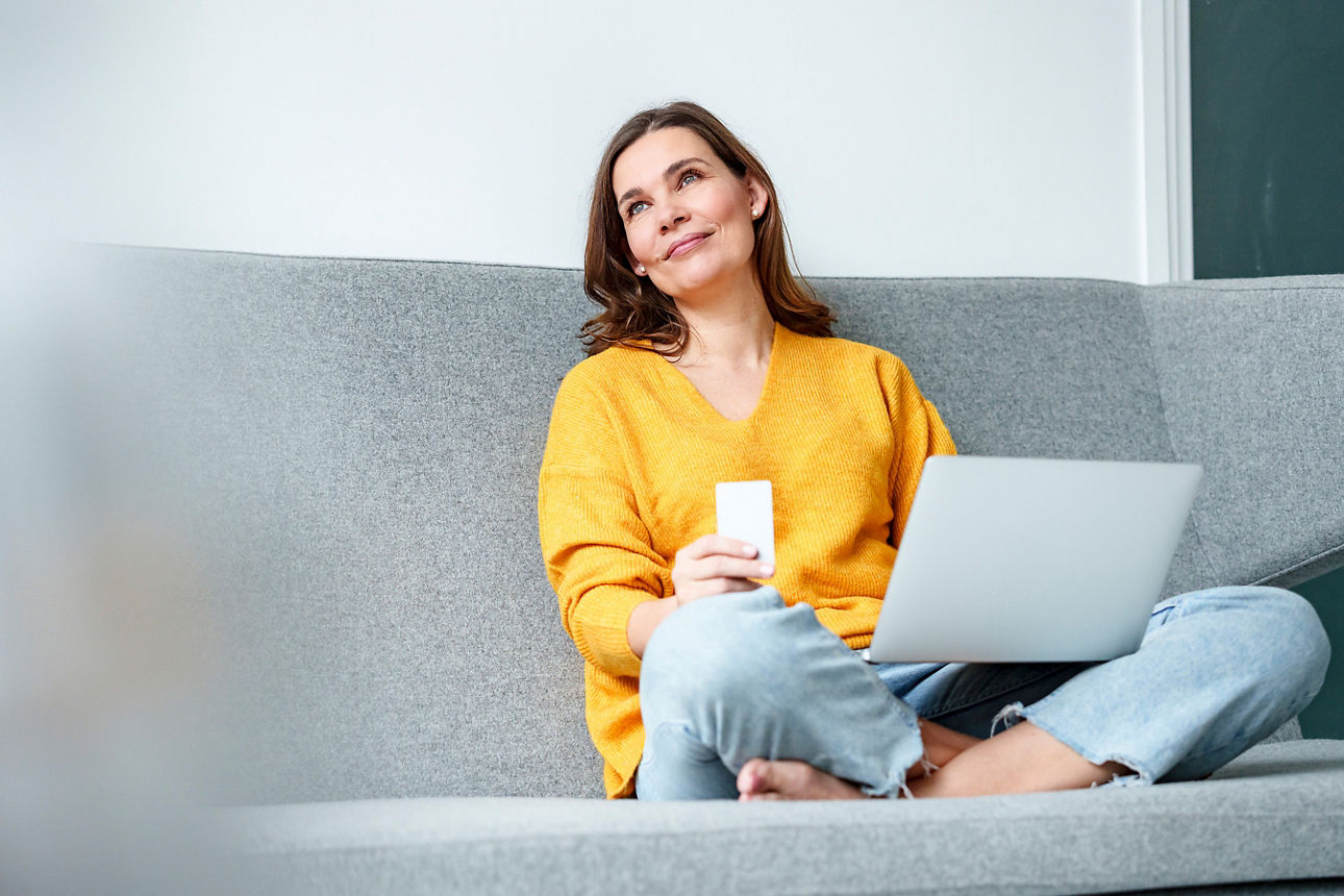 Eine Frau sitzt im Schneidersitz auf einer grauen Couch mit einem aufgeklappten Laptop auf ihrem Schoß