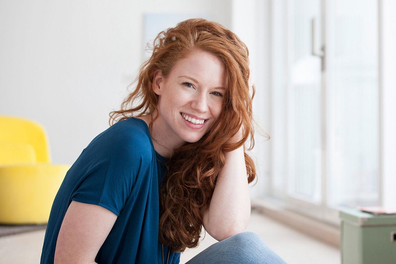 Eine lächelnde rothaarige Frau sitzt entspannt in einem hellen Raum mit gelbem Sessel im Hintergrund