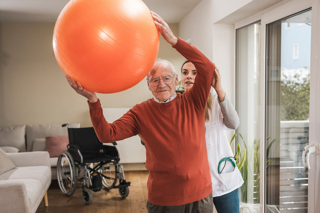 Hauspflegerin mit älterem Mann, der Übungen macht und einen Fitnessball hält
