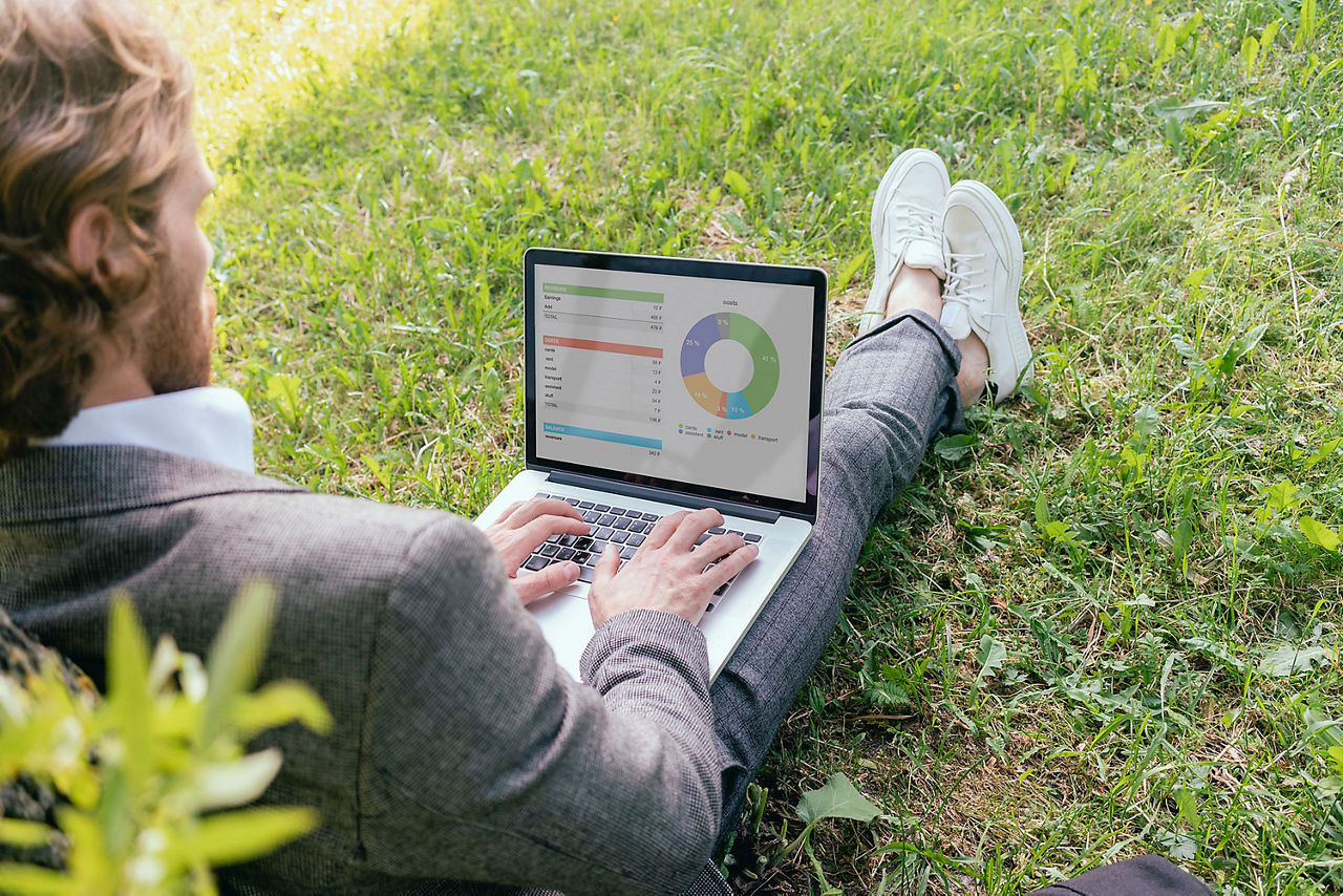 Ein Mann sitzt angelehnt an einem Baum mit aufgeklappten Laptop vor sich auf einer grünen Wiese.