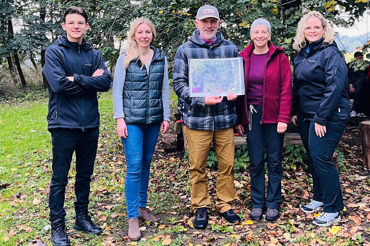 Fünf Erwachsene stehen draußen auf einer Lichtung und posieren gemeinsam für ein Gruppenfoto. Der Mann in der Mitte hält ein gerahmtes Bild mit einem Naturmotiv. Die Gruppe wirkt fröhlich und verbunden.