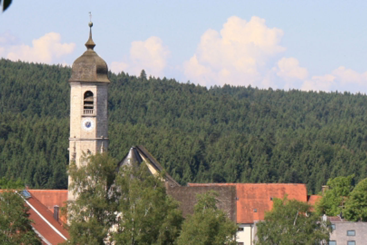 Panoramabild von Weyarn mit einer historischen Kirche mit hohem Zwiebelturm und einem modernen gelben Baukran im Vordergrund, umgeben von Wäldern und Häusern.