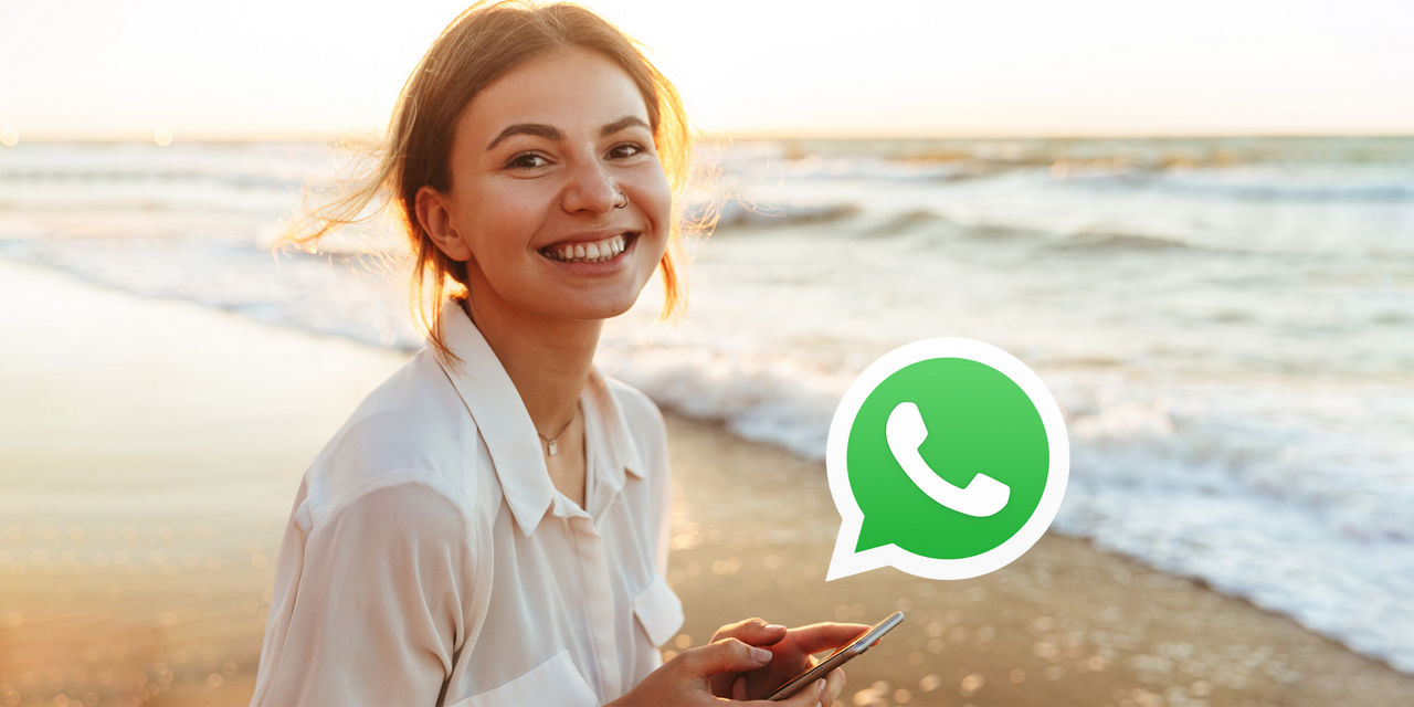Eine Person am Strand hält ein Smartphone, während das WhatsApp-Logo im Vordergrund sichtbar ist.