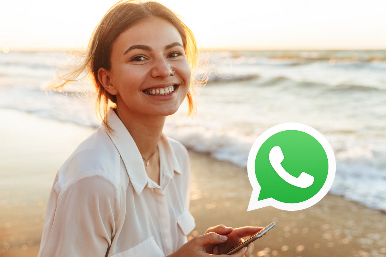 Eine Person am Strand hält ein Smartphone, während das WhatsApp-Logo im Vordergrund sichtbar ist.