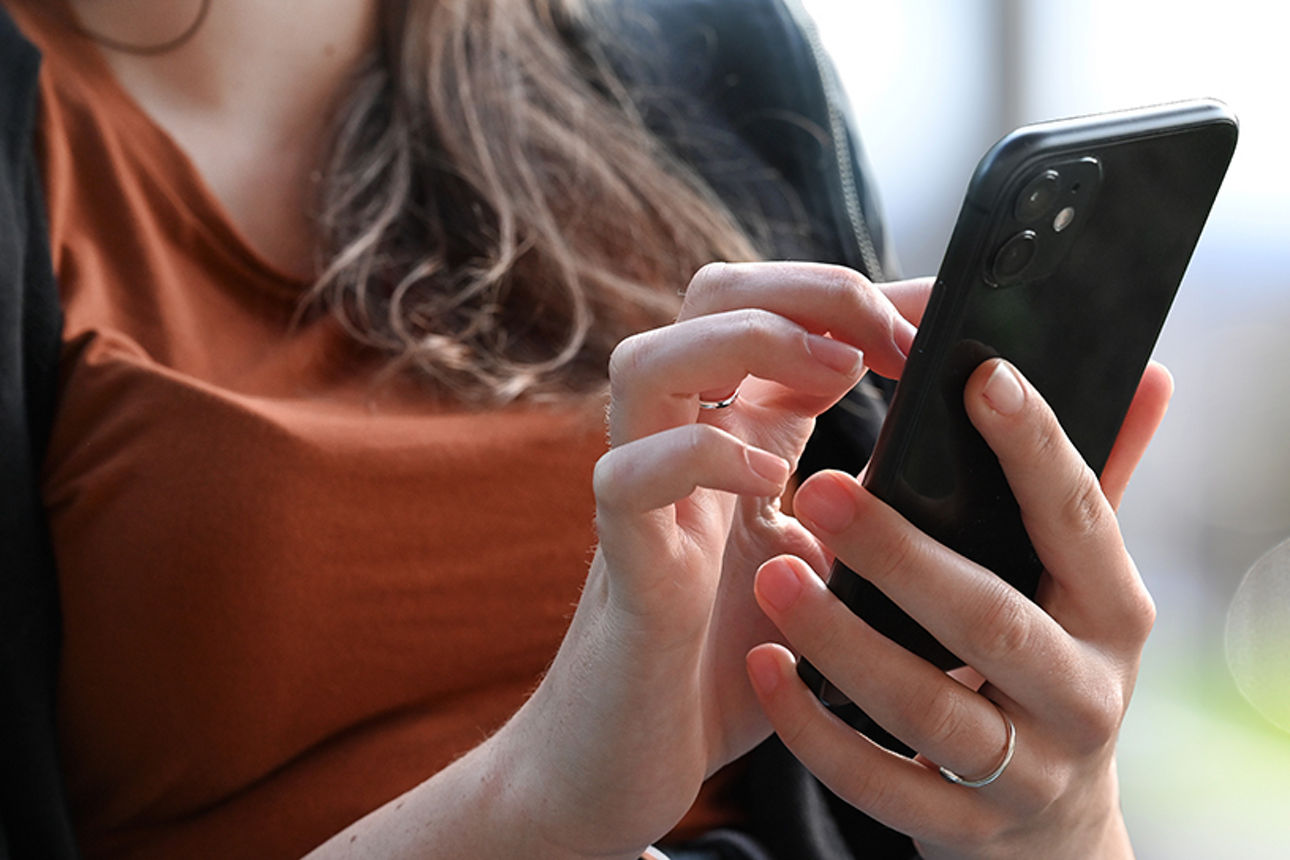 Frau mit braunem Pullover bedient ein Smartphone. Im Hintergrund sind unscharfe Grünflächen und ein Gebäude zu sehen.