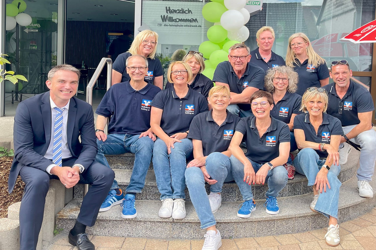 Einige Personen sitzen vor einer Bankfiliale und tragen ein Volksbank-Shirt. Links eine Person im Anzug.