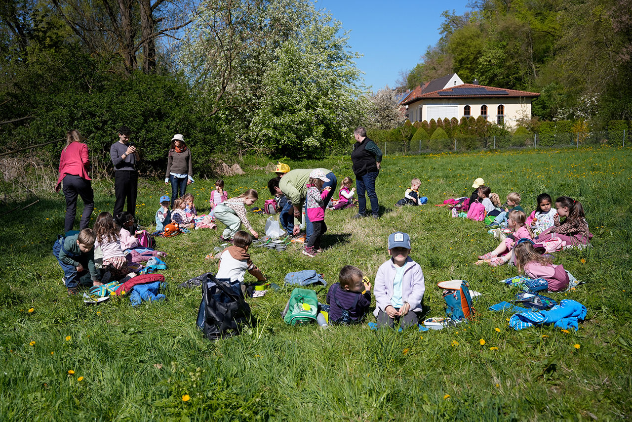 Gruppe von Kleinkindern mit 5 Erwachsenen macht Pause bei einem Ausflug auf einer Wiese.