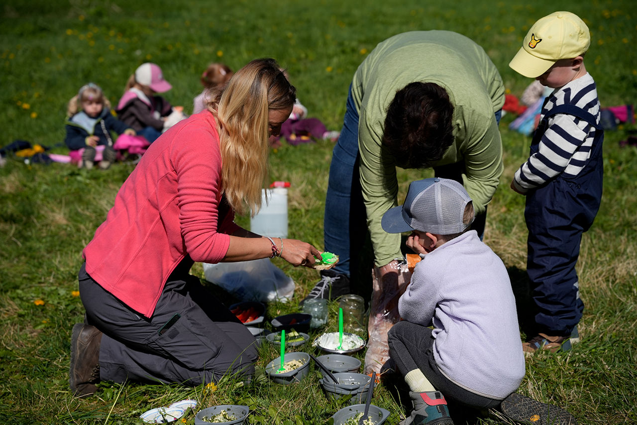 Zwei kleine Jungs mit Käppies und zwei Erwachsene machen Brotzeit auf einer Wiese. Im Hintergrund weitere Kinder