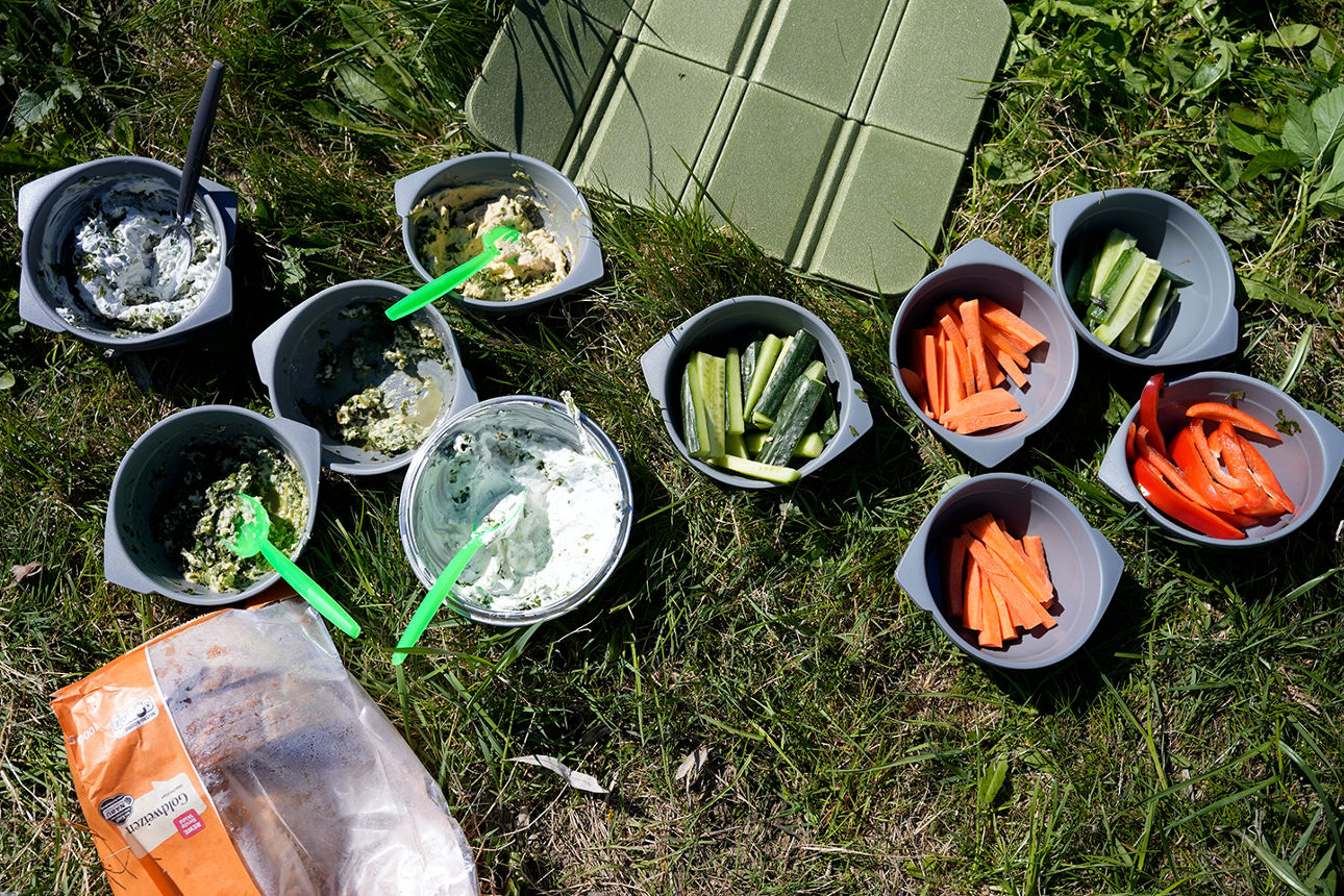 Auf einer Wiese stehen mehrere Plastikschüsseln mit aufgeschnittenem Gemüse und frisch angerührten Dipps