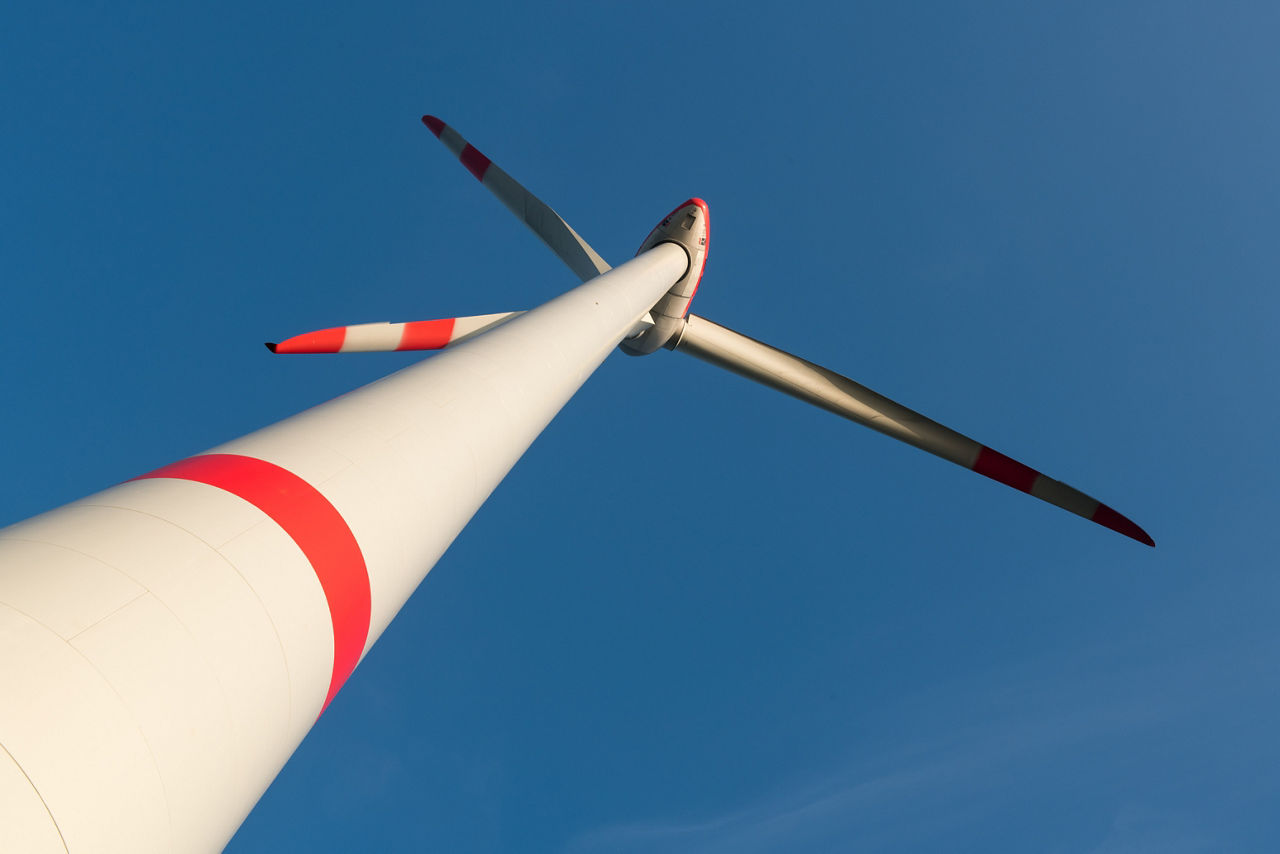 Nahaufnahme einer modernen Windkraftanlage mit rot-weiß-markierten Rotorblättern, vor einem klaren, blauen Himmel .