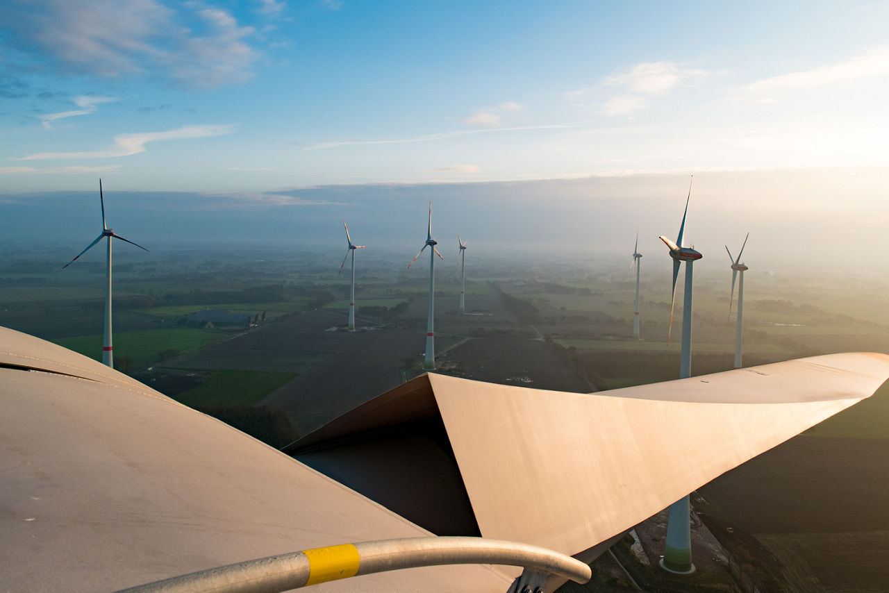 Luftaufnahme eines Windparks. Im Vordergrund ist ein Windradflügel zu sehen, im Hintergrund weitere Windräder..