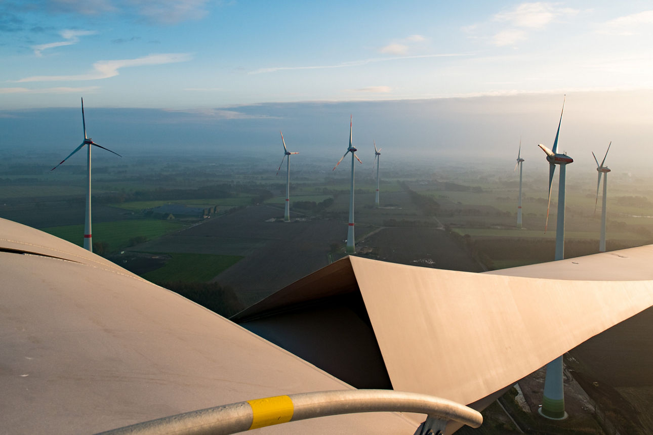 Luftaufnahme eines Windparks. Im Vordergrund ist ein Windradflügel zu sehen, im Hintergrund weitere Windräder..