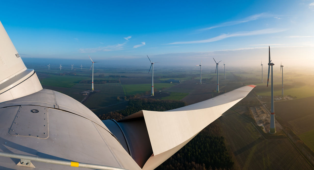 Luftaufnahme eines Windparks. Im Vordergrund ist ein Windradflügel zu sehen, im Hintergrund weitere Windräder.