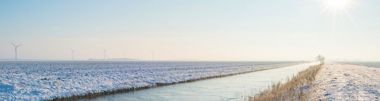 Sonne über einem gefrorenen sonnendurchfluteten Kanal mit Schnee und Mühlen im Hintergrund