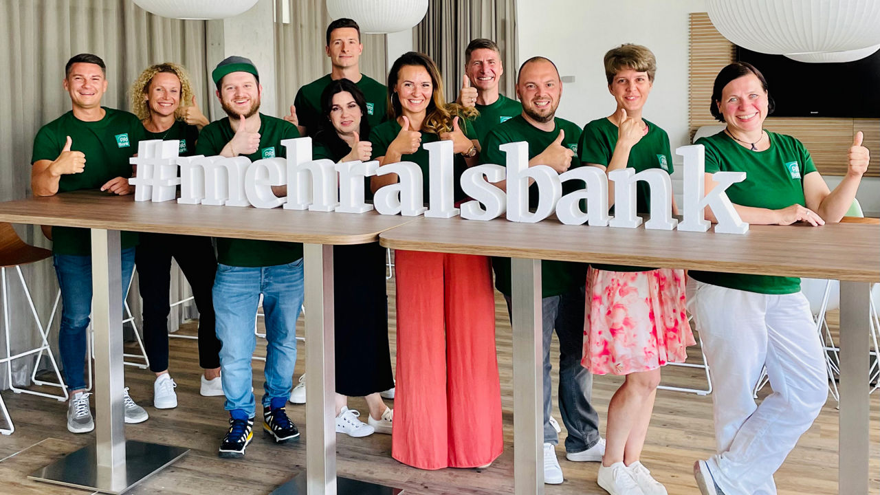 Mitarbeitende die lächelnd in die Kamera schauen und hinter dem Schriftzug #mehralsbank stehen.