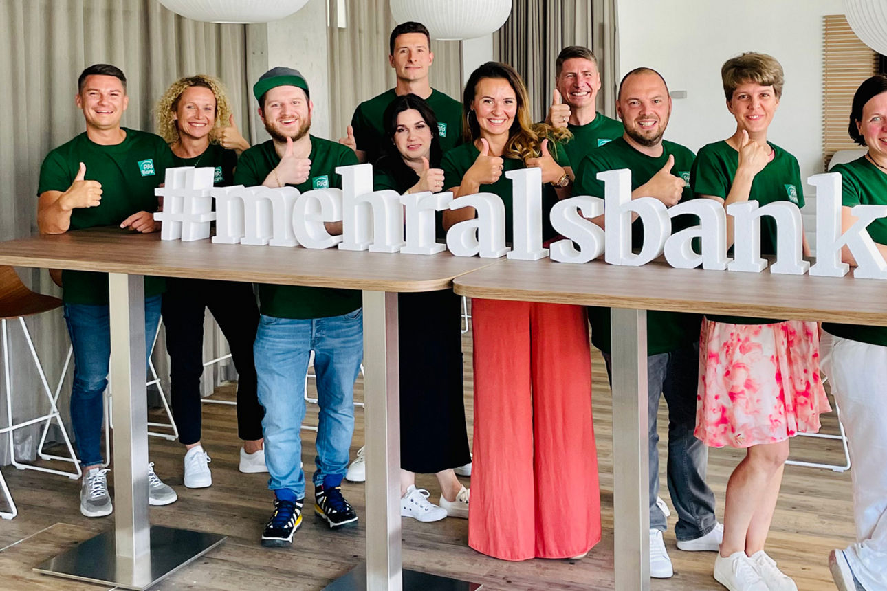 Mitarbeitende der PSD Bank stehen hinter der Schrift #mehralsbank und lächeln in die Kamera.