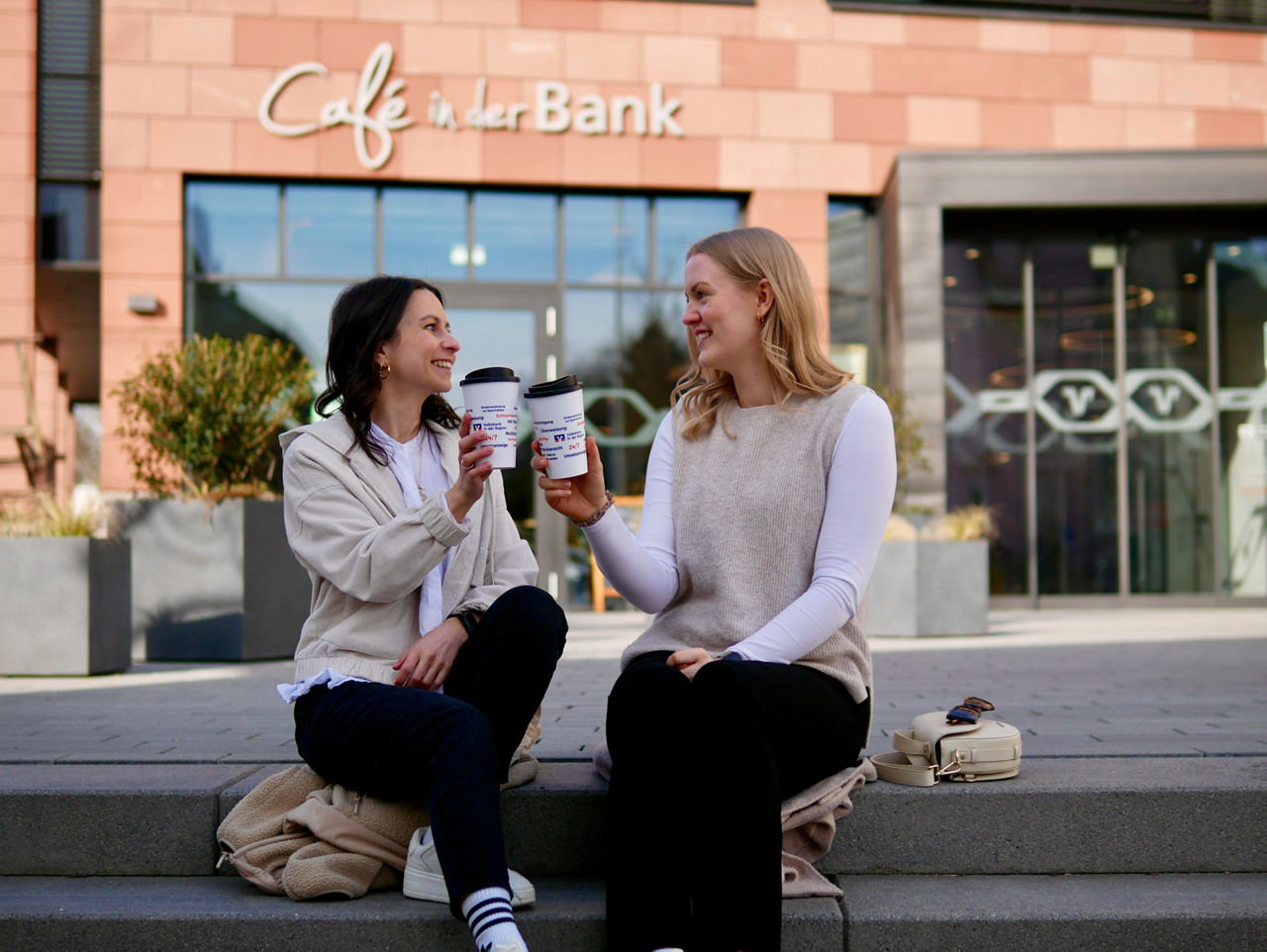 Zwei junge Frauen sitzen mit Kaffeebecher auf der Treppe vor der Volksbank