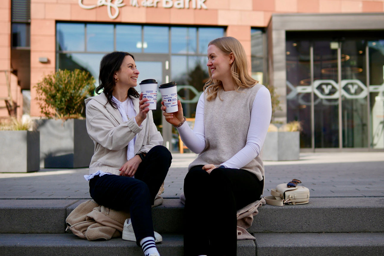 Zwei junge Frauen sitzen mit Kaffeebecher auf der Treppe vor der Volksbank