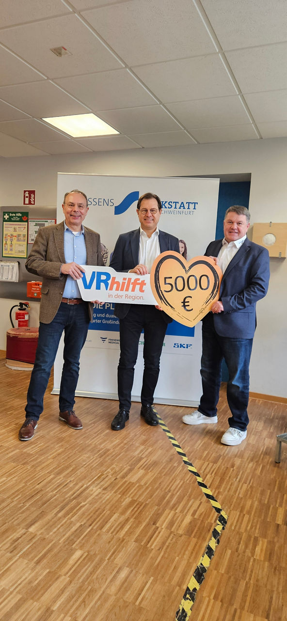 3 männliche Personen stehen in einem Raum. Sie halten ein großes, herzförmiges Schild mit der Aufschrift 5000 Euro. 