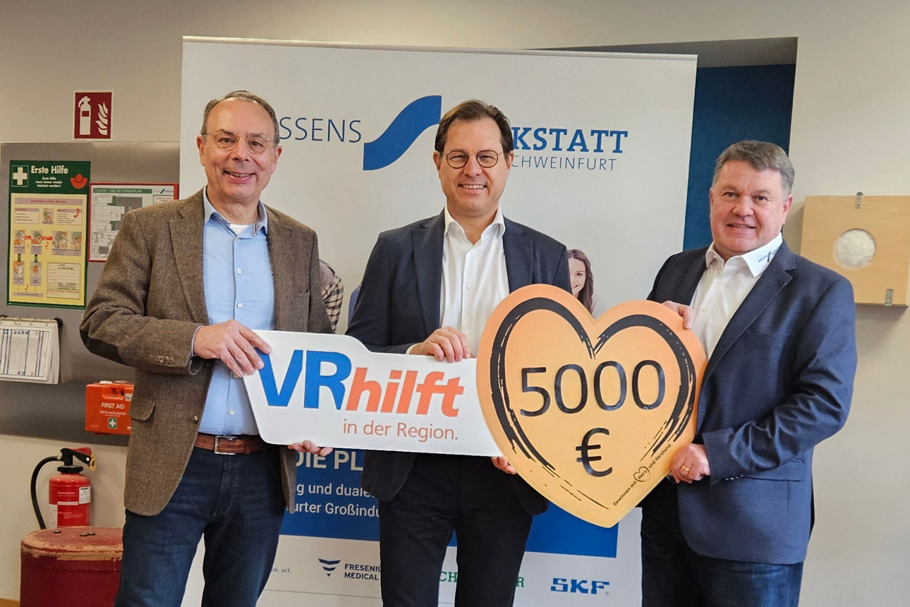 3 männliche Personen stehen in einem Raum. Sie halten ein großes, herzförmiges Schild mit der Aufschrift 5000 Euro. 