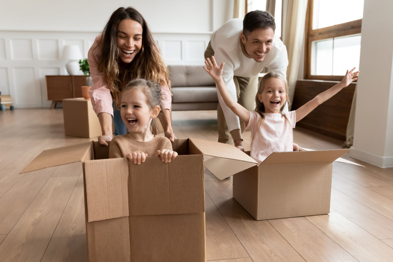 Fröhliche Mädchen sitzen während eines Rennwettbewerbs mit den Eltern in großen Kartons in ihrem neuen modernen Zuhause am Umzugstag. 