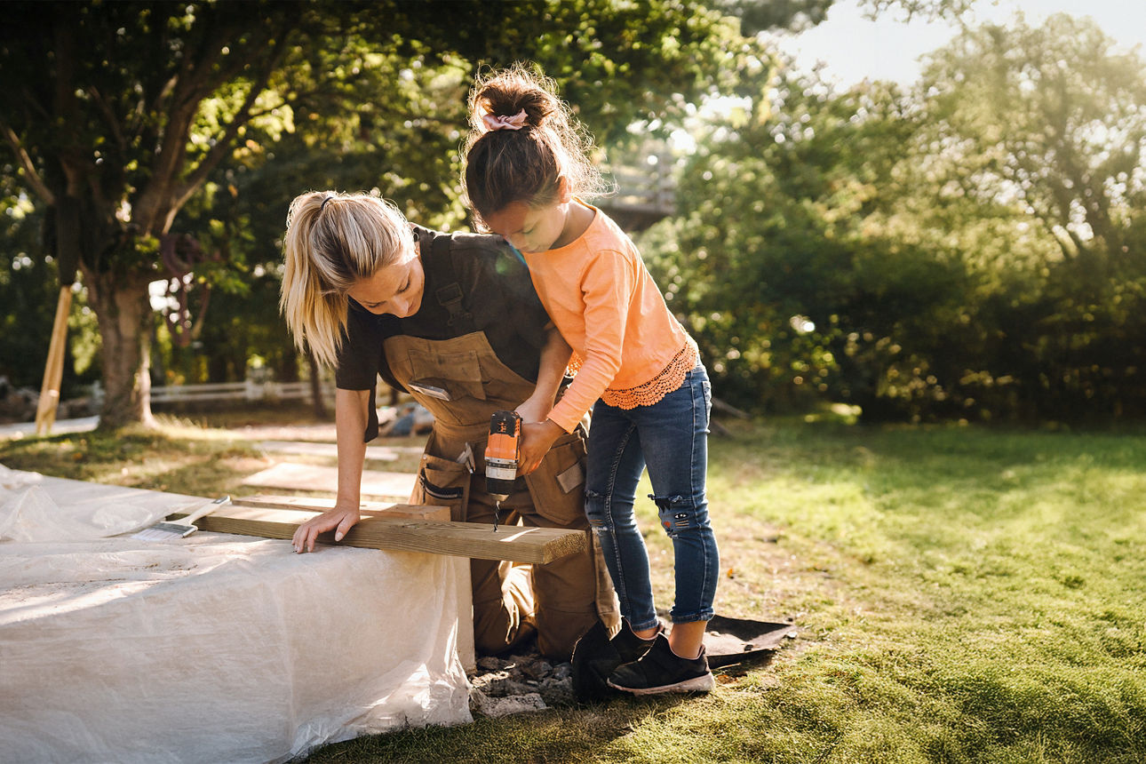 Ein Mädchen sägt unter Aufsicht der Mutter ein Holzbrett durch.
