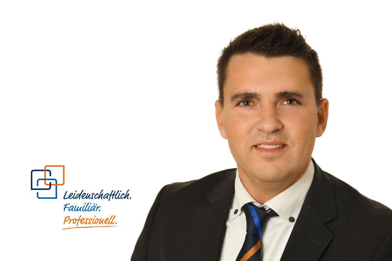 Wolfgang Hampel, ein junger Mann mit kurzen, dunklen Haaren, lächelt freundlich und trägt einen dunklen Anzug mit einer gestreiften blau-orangenen Krawatte.