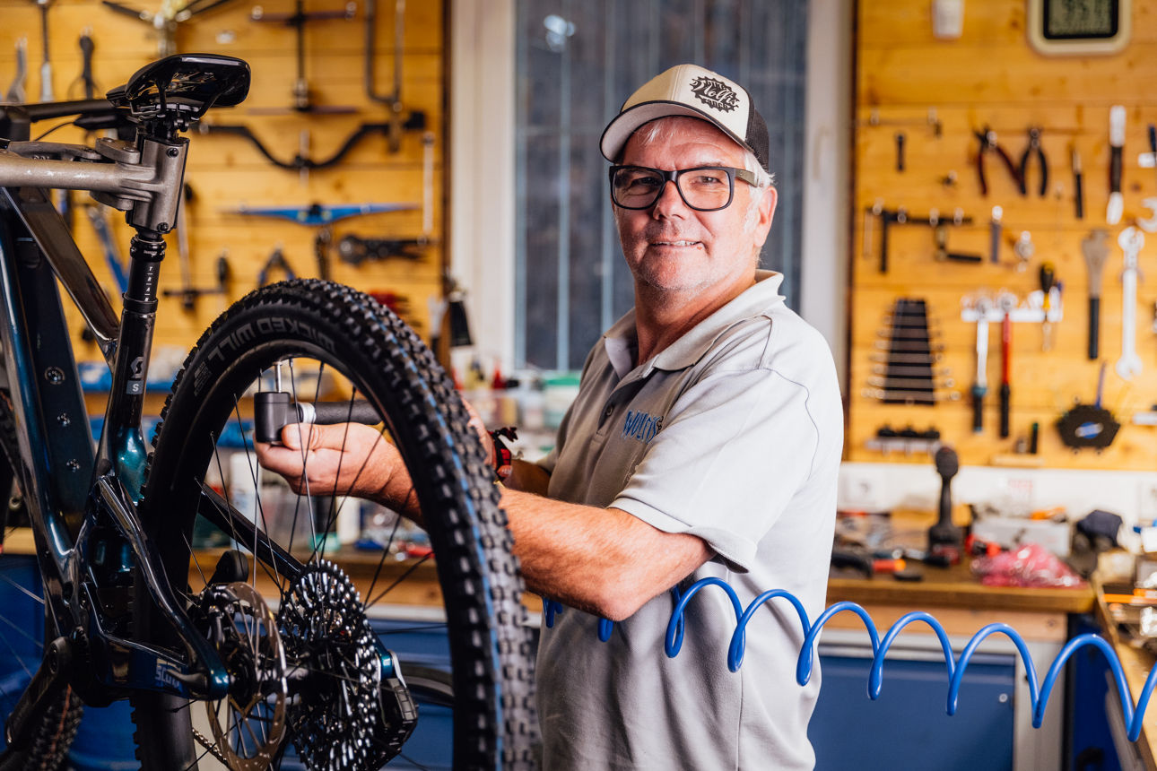 Innenaufnahme einer Fahrradwerkstatt bei Wolfis Bike Ranch mit einem Fahrrad auf Montageständer. Eine Person arbeitet im Vordergrund an der Technik, im Hintergrund sind Werkzeugwände und Arbeitsmaterialien sichtbar.