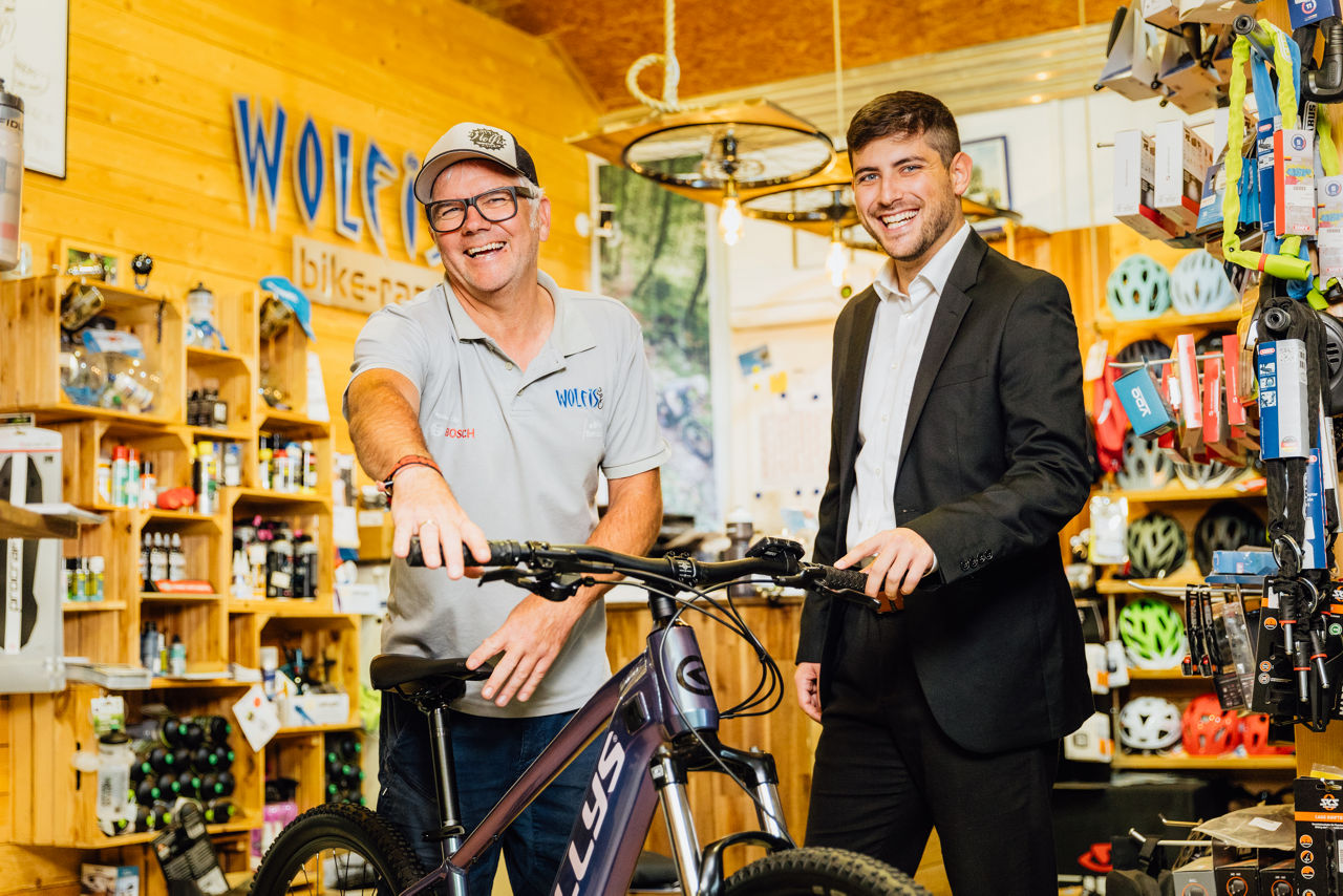 Wolfgang Brand und Berater Darius Sarfaraz stehen im Verkaufsraum von Wolfis Bike Ranch neben einem präsentierten Fahrrad. Die Szene zeigt den Austausch im Rahmen der Produktvorstellung zwischen Fahrrädern und Zubehör.