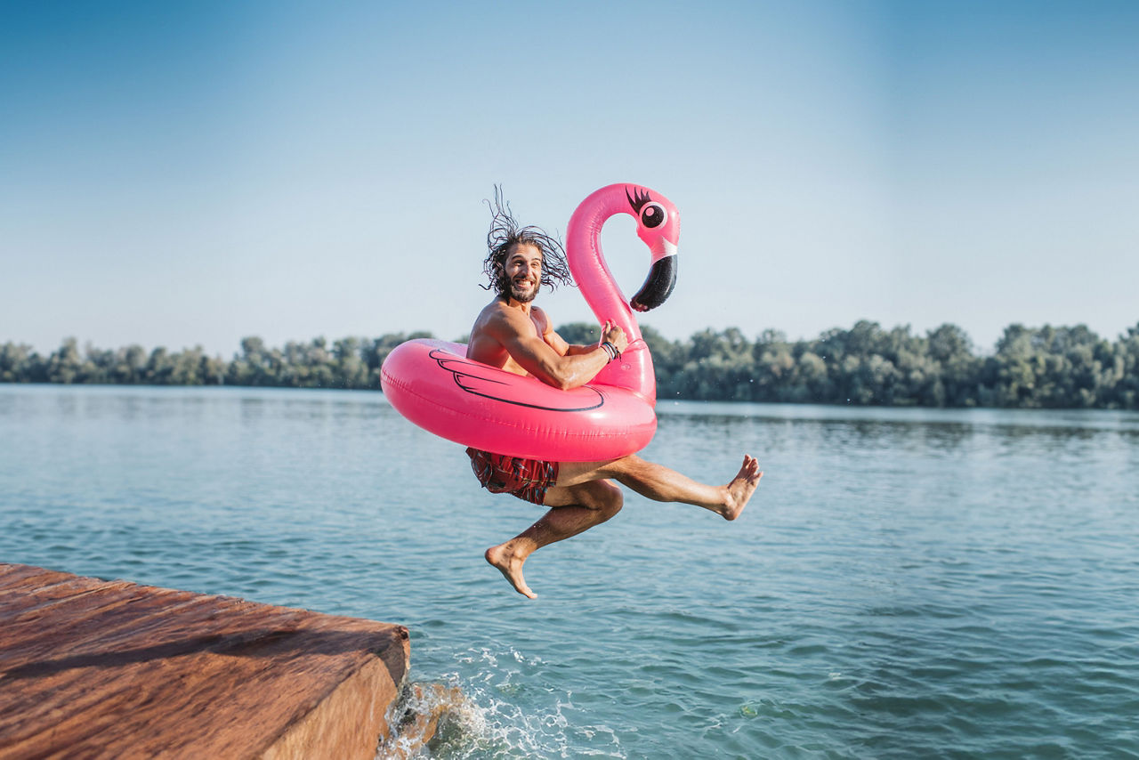 Ein junger Mann springt lachend mit einem pinken Flamingo-Schwimmring von einem Holzsteg in einen See.