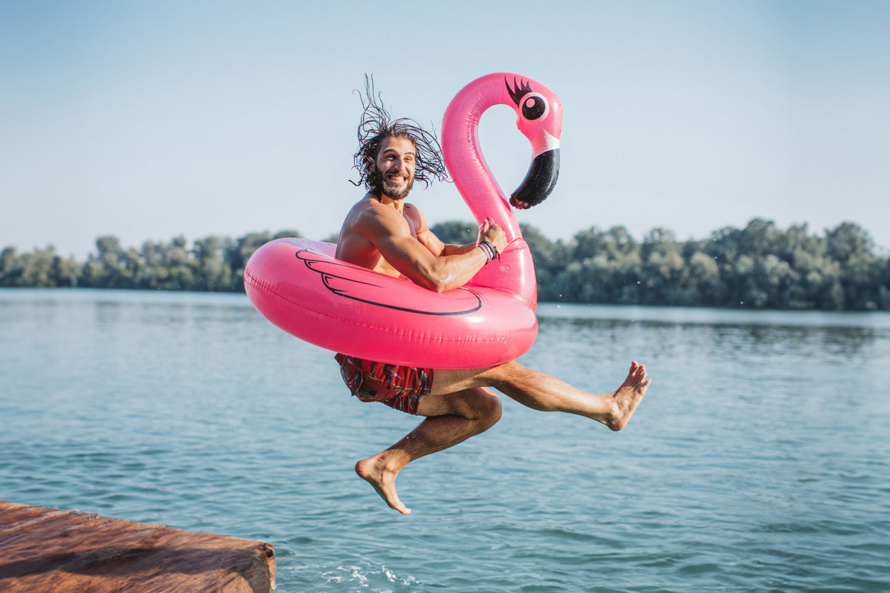 Ein junger Mann springt lachend mit einem pinken Flamingo-Schwimmring von einem Holzsteg in einen See.