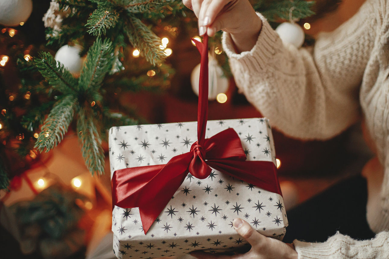 Hände in einem gemütlichen Pullover öffnen ein Weihnachtsgeschenk mit roter Schleife vor dem Hintergrund eines Weihnachtsbaums mit Lichtern. Stilvolle Frau hält ein Geschenk mit rotem Band in einem festlich dekorierten Raum aus nächster Nähe.