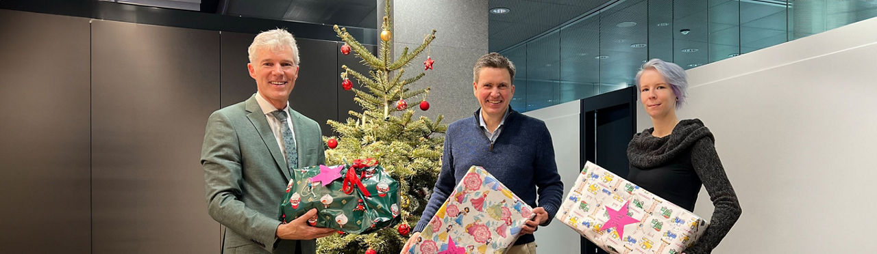 Drei Personen stehen vor einem Tannenbaum mit drei Geschenken in den Händen