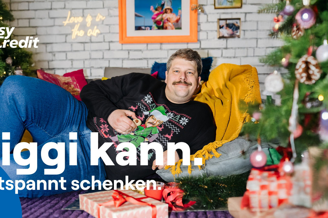 Siggi von der TeamBank liegt auf einer Couch und trägt einen Weihnachtspullover
