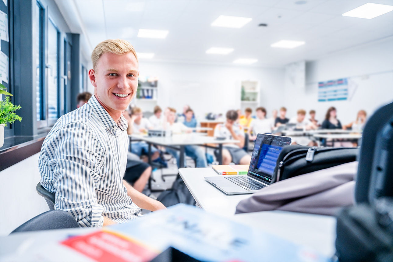 Person sitzt vor einer Schulklasse und lächelt in die Kamera