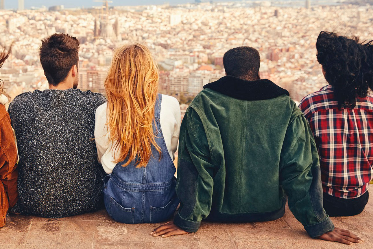 Rear view of friends sitting on building terrace. Males and females are looking at cityscape and sea. They are wearing casuals.