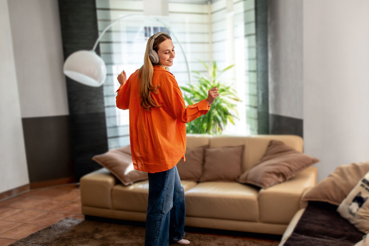 Eine junge Frau in Jeans und orangener Bluse tanzt durch ein Wohnzimmer.