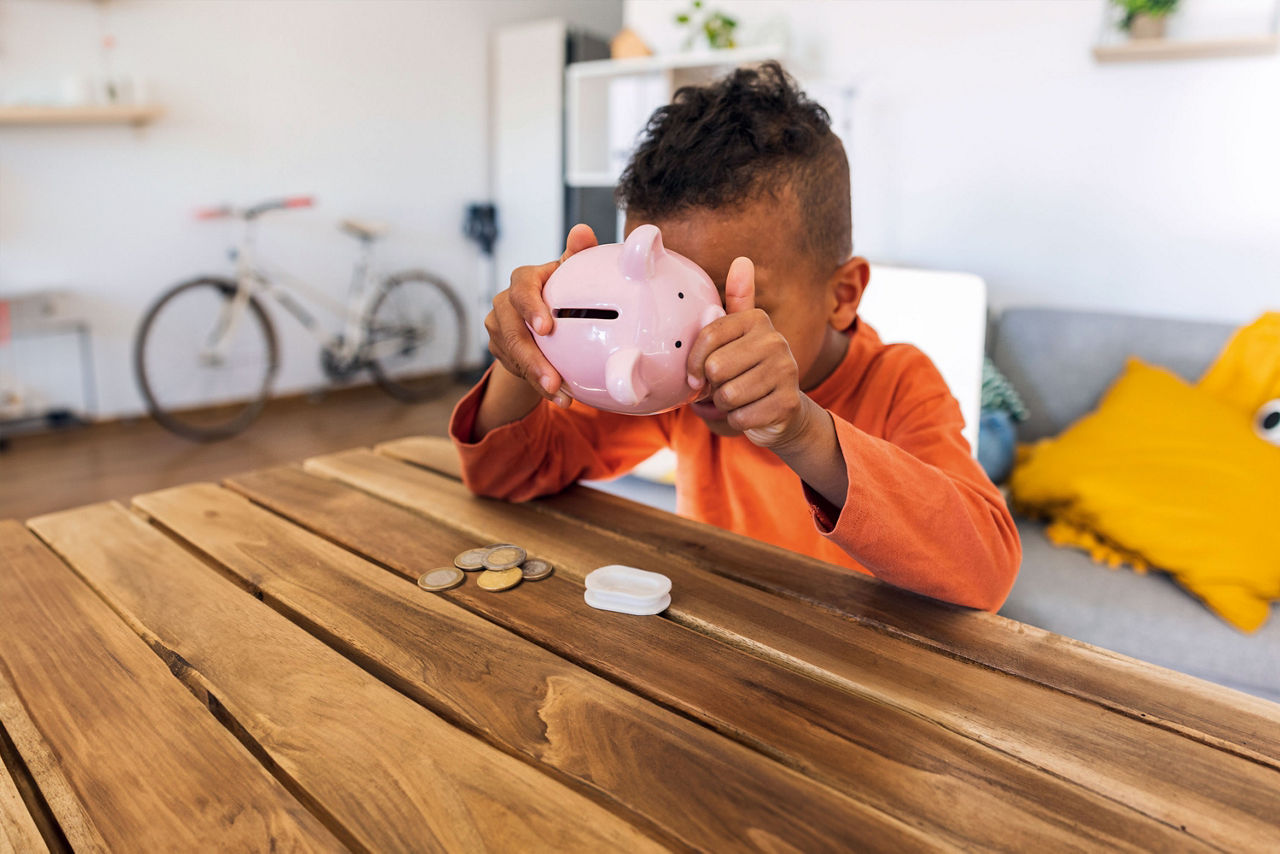 Junge mit orangenem Pullover sitzt am Tisch und leert sein rosanes Sparschwein