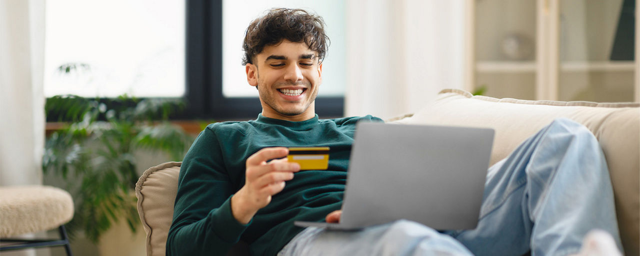 Ein junger Mann sitzt vor seinem Laptop und hält eine Karte in der Hand. Er lächelt.
