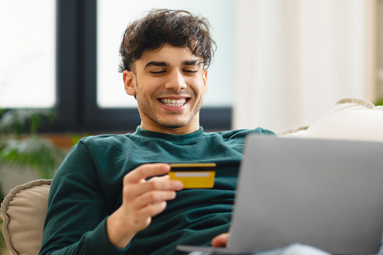 Ein junger Mann sitzt vor seinem Laptop und hält eine Karte in der Hand. Er lächelt.