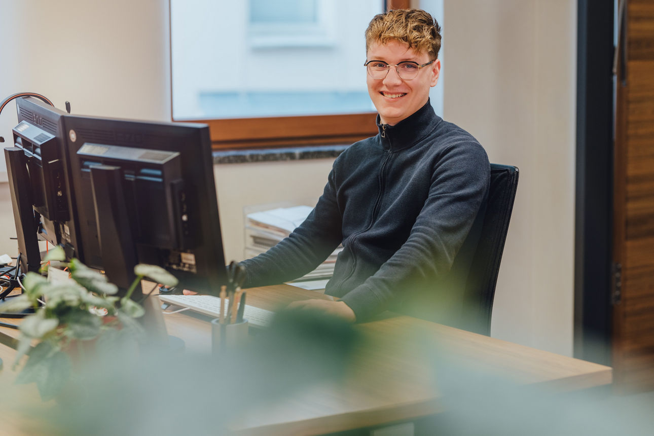 Ein Mitarbeiter sitzt vor dem Computer und lacht in die Kamera