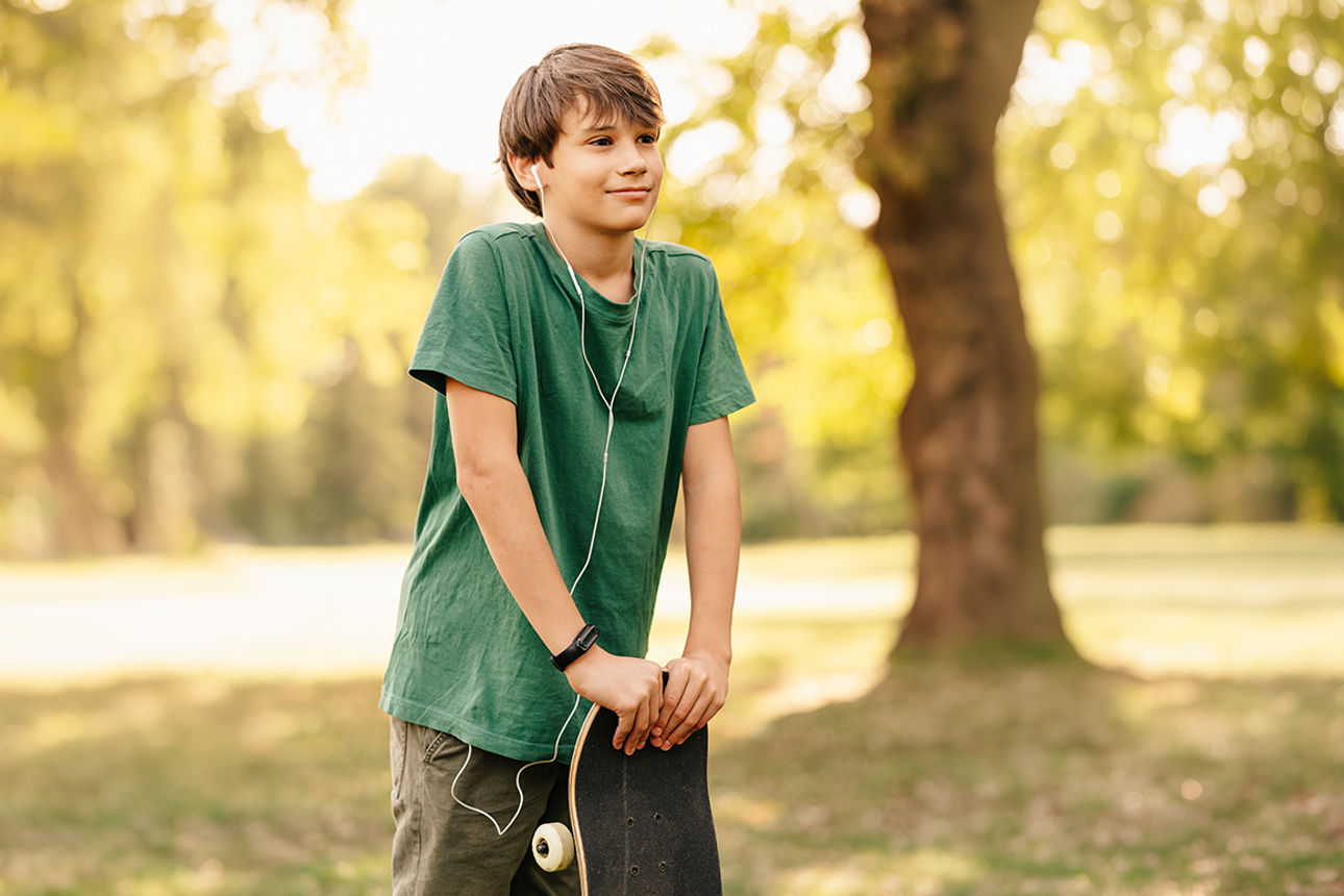 Ein Junge, der mit seinem Skateboard und Kopfhörern in der Natur steht.
