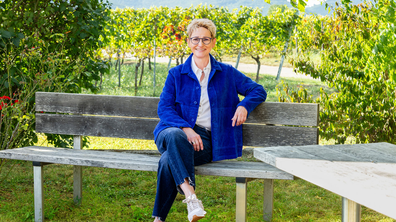 Lächelnde Frau mit Brille und weißer Bluse sitzt vor Weinreben im Freien.