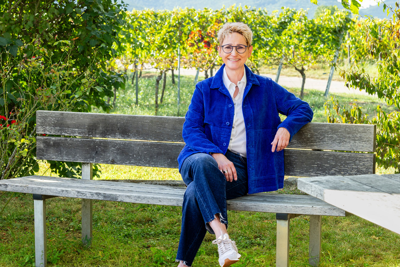 Lächelnde Frau mit Brille und weißer Bluse sitzt vor Weinreben im Freien.