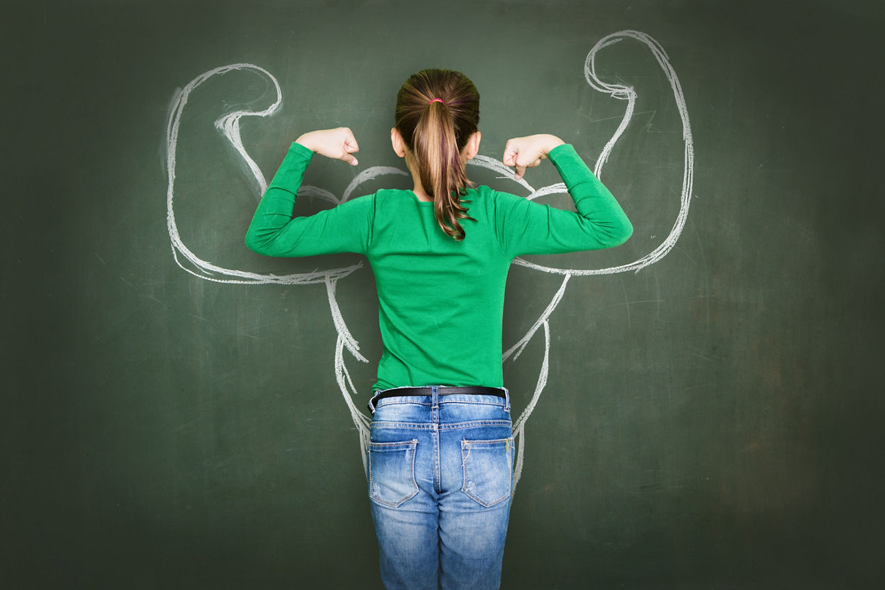 Ein Mädchen steht mit einer Bizeps-Pose mit dem Gesicht zur Tafel, auf dem mit Kreide eine große Bizeps-Pose aufgemalt ist.
