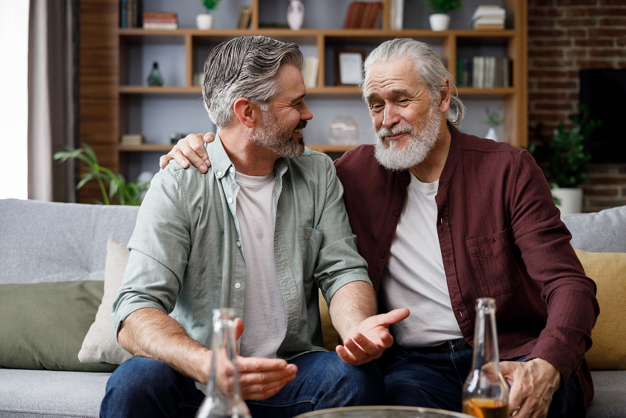 Alter Vater und reifer Sohn unterhalten sich auf dem Sofa sitzend und der Vater legt seinen Arm um den Sohn 