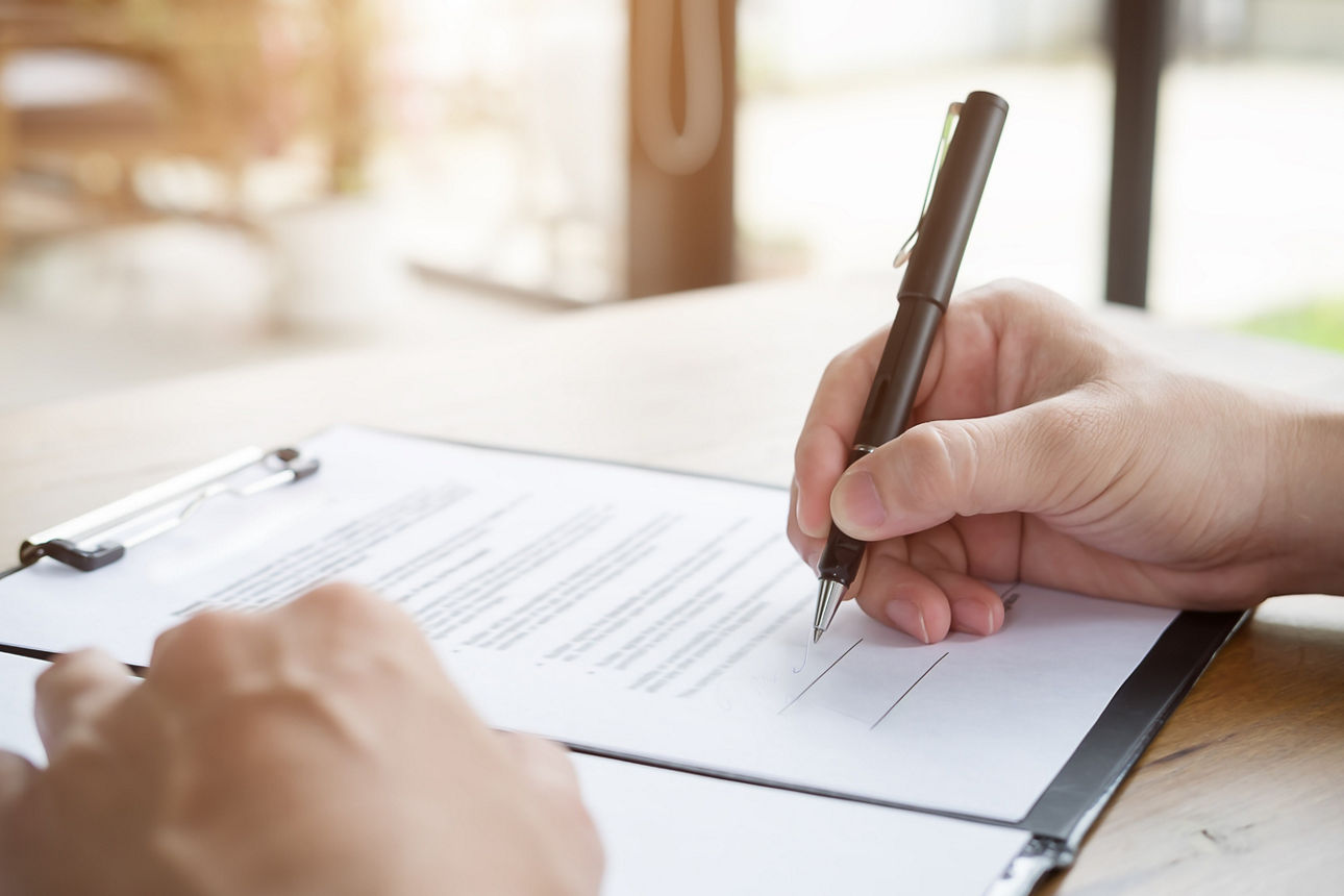 Eine Hand unterschreibt ein auf einem Tisch in eine Mappe liegendes Dokument