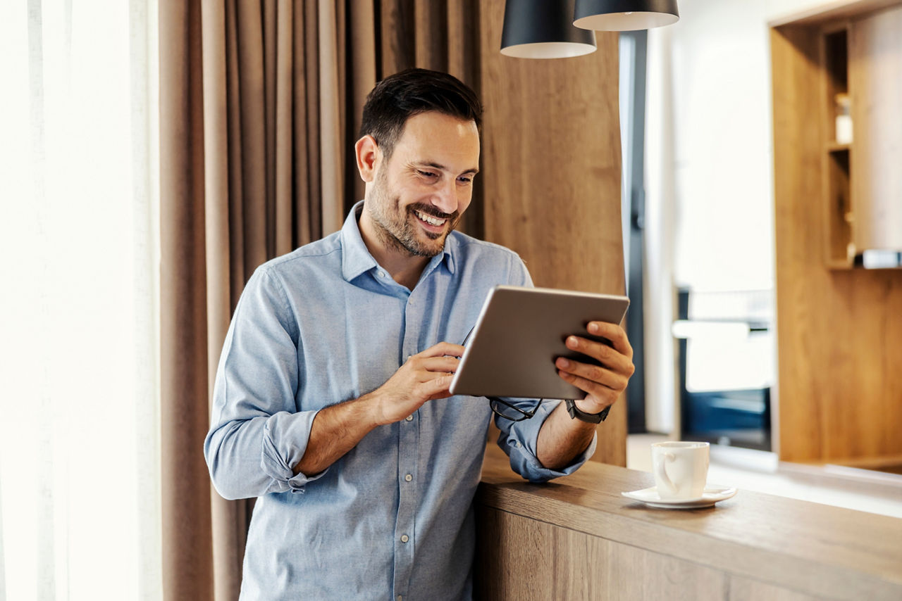 A man stands at home and reads e-mails on tablet while smiling at display.; Shutterstock ID 2280974995; purchase_order: Purchase Order: Website Atruvia Bilder; job: VTM Bildauswahl; client: ; other: 